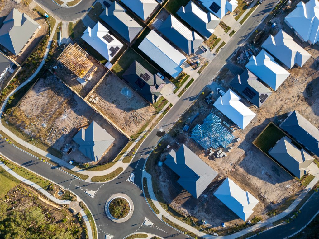 Top-down aerial view of a new Australian residential land release with completed homes, homes under construction at frame and slab stages, vacant lots, and a roundabout, showing multiple stages of residential development simultaneously.