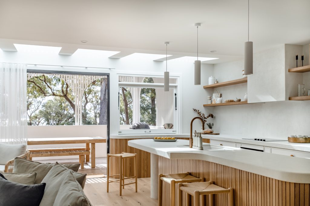 A bright, open-plan kitchen and living area in a renovated Australian home, featuring timber accents, a curved island bench, skylights, and large windows looking out to native eucalyptus trees.